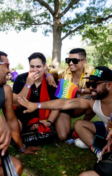 A group of people sit closely together on the grass in a shady park, interacting with one another during a Pride event, with trees overhead and a small rainbow Pride flag visible among the group.