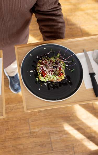 A person holds two wooden trays with black plates featuring colorful salads, garnished with microgreens, on a wooden floor.