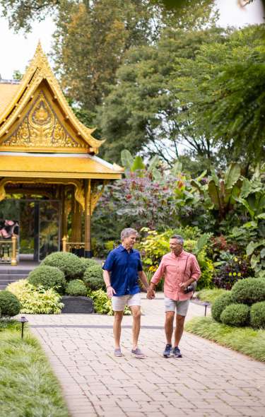Two men holding hands walk away from the gold Thai pavilion building at Olbirch Botanical Gardens. The couple is smiling at each other and is surrounded by lush greenery.