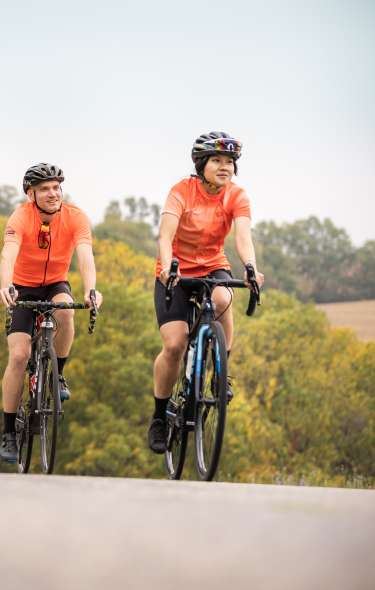 Two men biking up hill wearing bright orange shirts, helmets, and biking shorts. The trees in the background show leaves just beginning to change color for Fall.