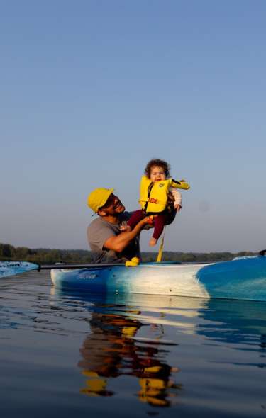 A man holds up a young toddler who is wearing a life vest as they float in a canoe on Lake Wingra