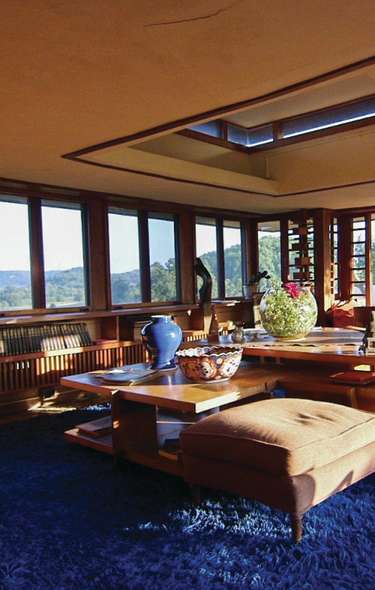 The living room in one of Frank Lloyd Wrights iconic buildings. The room has low ceilings and is lined with book shelves and large windows looking out onto a patio and lush wooded scenery. Inside there is blue shag carpeting underneath low tables and seating arrangements