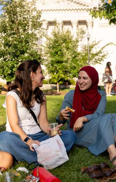 Two women, one wearing a hijab, sit and have a picnic on the lawn in front of the Capitol building