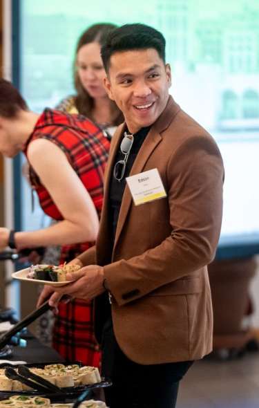 A man smiles at a person out of frame as he adds food to his plate at a buffet line at a meeting.
