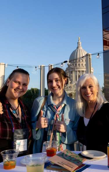 Three white women conference attendees share drinks and thumbs of the camera on the rooftop of the Madison Children's Museum with the State Capitol building in the background