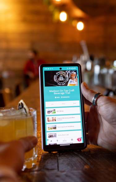 A person with paw print tattoos on their arm holds a phone with the Madison On Tap pass showing on the screen in one hand and a cocktail in the other hand