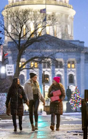 A trio of women walk in the snow in downtown Madison carrying presents with lights around them and the Wisconsin State Capitol building glowing behind them