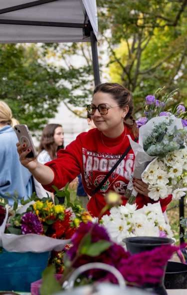 A woman takes a photo on her cell phone while holding bouquets of flowers at the Dane County Farmers' Market