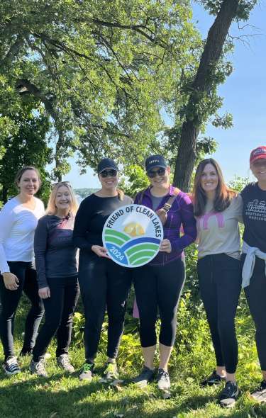 A group of six white women stand on the lakeshore in athletic clothing that says "Friends of Clean Lakes 2024."