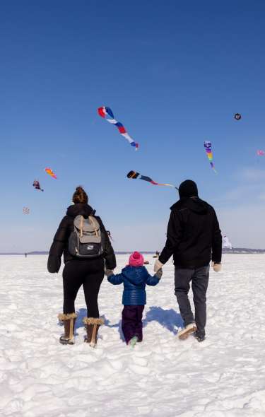 Two adults hold a small child's hand while walking on a frozen lake and looking at a sky full of kites.