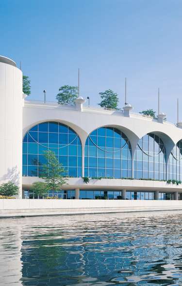 A view of Monona Terrace from Lake Monona. The sky is clear and sunny and a reflection of the building at its larger arched windows can be seen in the lake water.