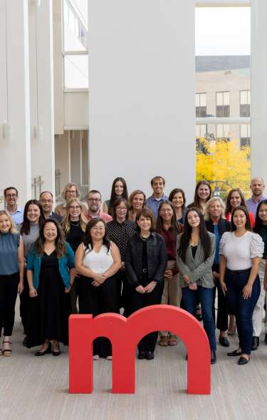 The entire Destination Madison team of about 35 people stand in a group at the Overture Center with a large red m statue in front of them