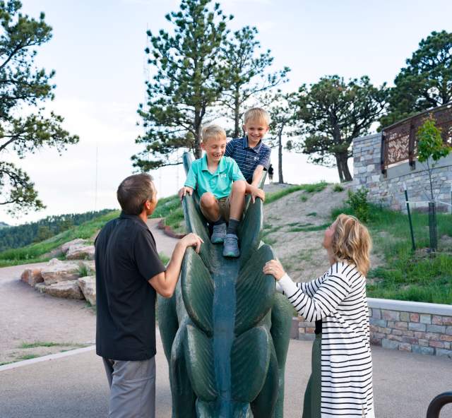 Two grinning boys sit on a green concrete dinosaur while looking at their parents standing on the ground with their hands on the dinosaur's back.