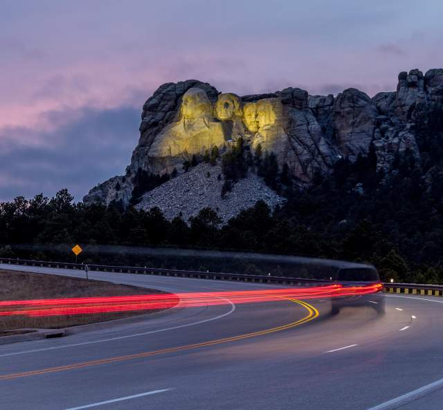 Mount Rushmore, lit up by yellow floodlights at dusk, sits in the background as a vehicle traveling down the highway is trailed by a long line of red taillights, created by a long-exposure photo.