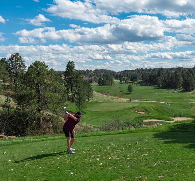 A man dressed in a black polo and black shorts tees off at the Golf Club at Red Rocks. Pine trees, a perfectly manicured course, bunkers, and a partly cloudy sky are visible in the distance.