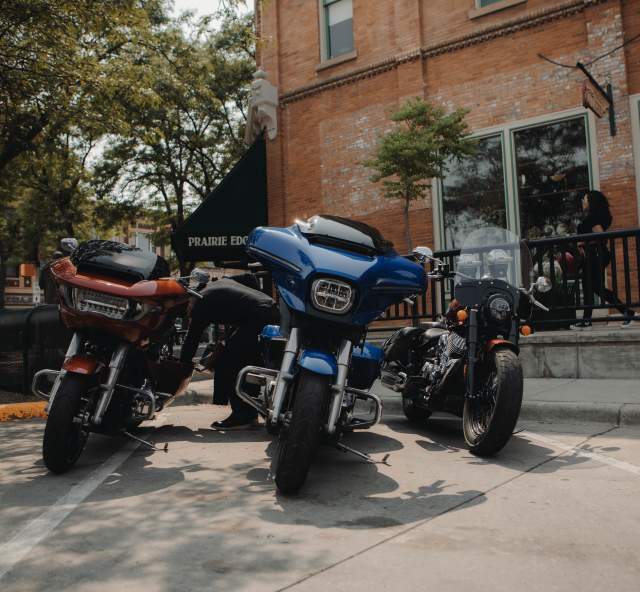 Three motorcycles, one red, one blue, and one black, are parked along the sidewalk, partially shaded by a tree, outside of Prairie Edge in Downtown Rapid City in mid afternoon.