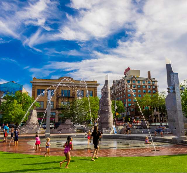 Children running through a splash patio next to a patch of green and surrounded by charming buildings. Blue sky with clouds.