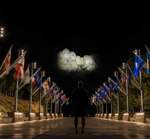 person standing at the avenue of flags looking up at an illuminated mount rushmore in the black hills of south dakota