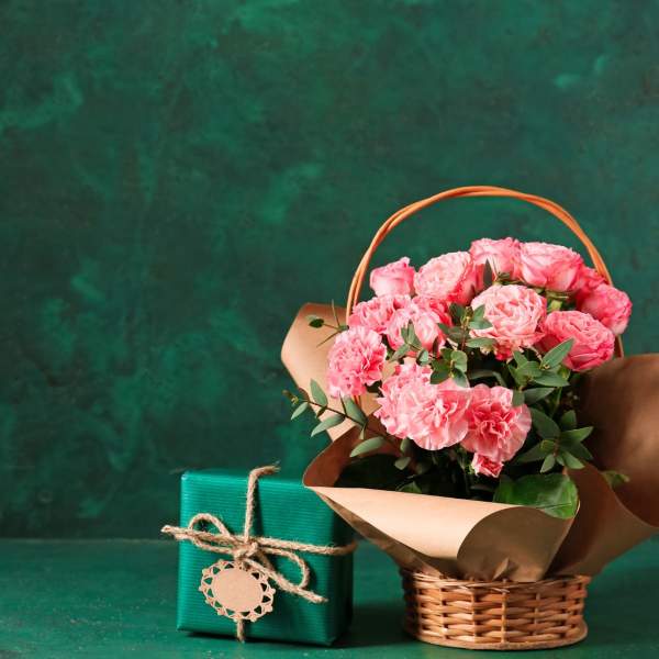 A wicker basket filled with pretty pink flowers next to a small square parcel wrapped in green paper and string with a gift tag