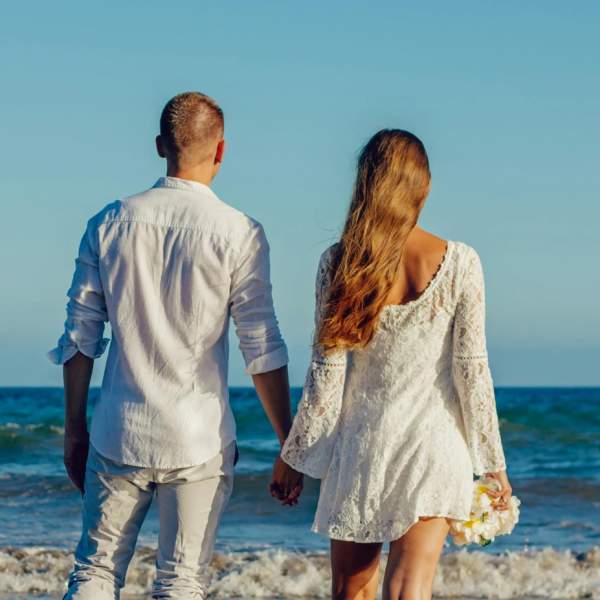 Photograph showing a couple dressed in white from the back as they stand on a beach