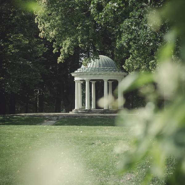 Den hvite og grønne paviljongen i parken på Hafslund Hovegård, sett gjennom bladverket til en busk.