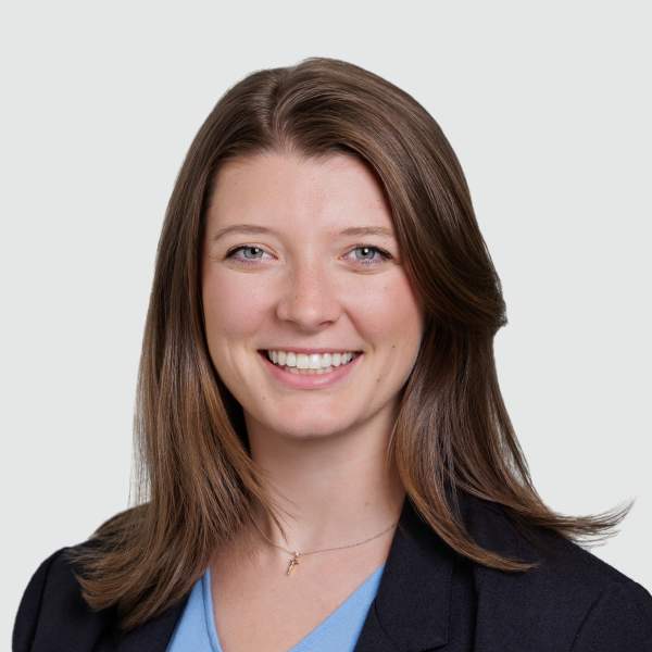 Woman with brunette hair, smiling, wearing a dark blue blazer and light blue shirt.