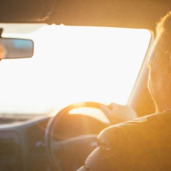 Photograph showing a man driving a car, as viewed from the back seats. The light is of the evening's golden hour.