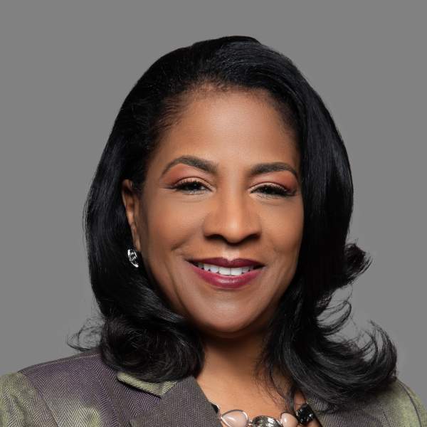 Professional headshot of a woman with black hair, smiling, wearing a grey blazer and brown shirt.