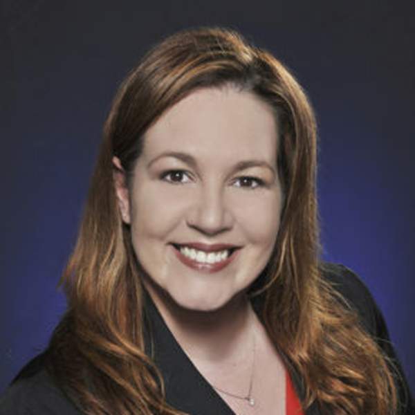 Professional headshot of a woman with long auburn hair wearing a black blazer over a red top, smiling against a dark gradient background.