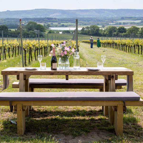 A bottle of English sparkling wine and flowers on a picnic bench at Roebuck Estate vineyard, with vines in the distance