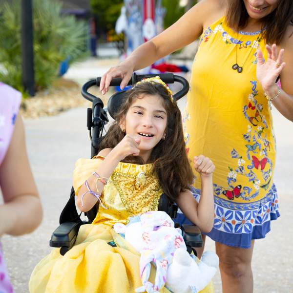 A young girl in a yellow princess dress smiles while sitting in a wheelchair, accompanied by a woman in a matching yellow outfit, as they enjoy a day outdoors.