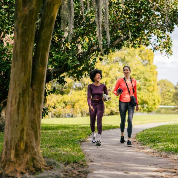 Two women in workout gear carry yoga mats under their arms as they stroll beneath oak trees in Orlando's Loch Haven Park