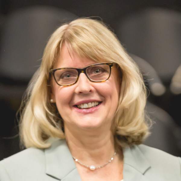 Professional headshot of a woman, with blonde hair, black glasses, smiling, with a sage green blazer.
