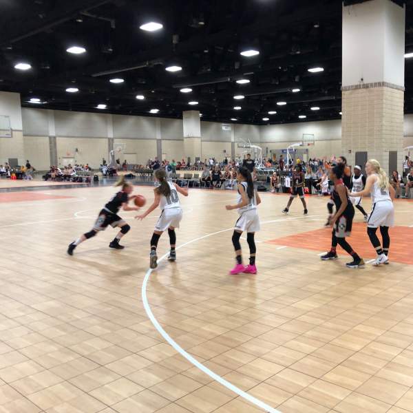 A high-energy basketball game unfolds in a large indoor arena, with players in white, black, and pink jerseys competing for the ball on a wooden court.