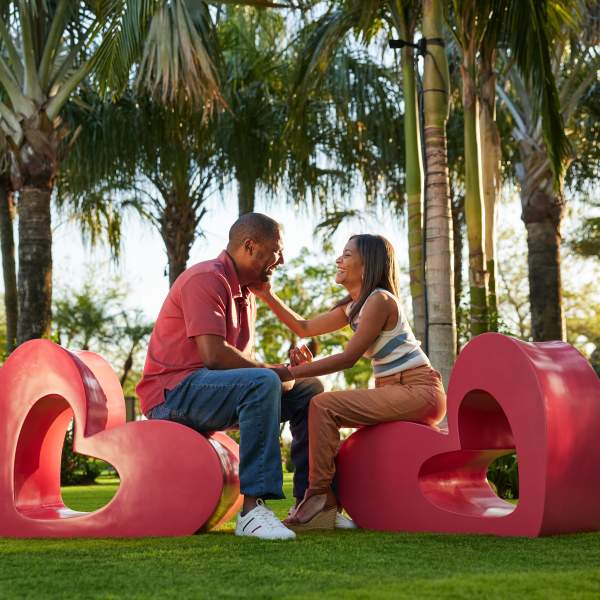 A couple sit facing each other on two red, heart-shaped seats, green grass at their feet and palm trees in the background