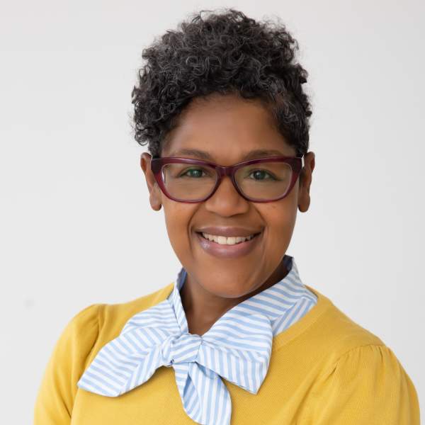 A black woman in a yellow sweater and glasses smiling at th ecamera.