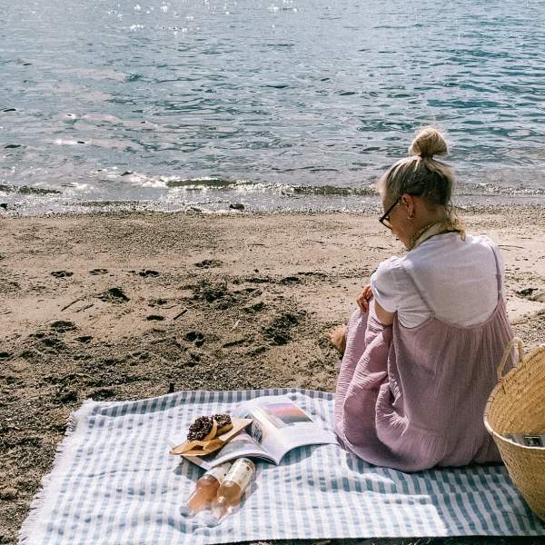 Kvinne som sitter på stranden på et badehåndkle med mat og drikke. Foto.