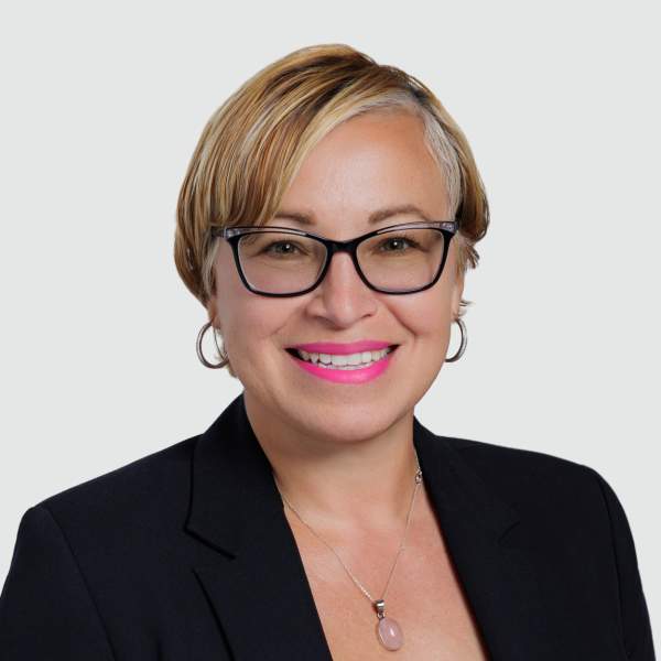 Woman smiling wearing pink lipstick, dark eyeglass frames, in black blazer.
