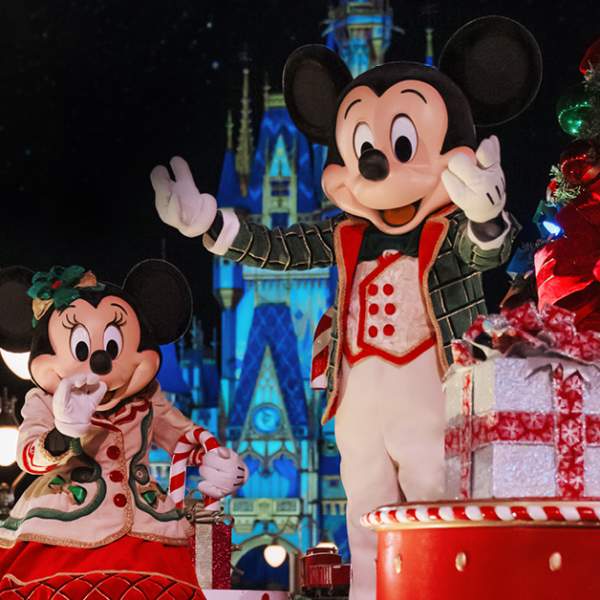 Mickey Mouse and Minnie Mouse riding a holiday parade float while wearing red, white and green holiday outfits. Cinderella Castle is visible in the background.