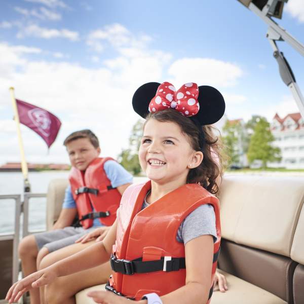 A girl wearing a life vest and mouse ears smiles as she and her brother ride past Disney's Grand Floridian Resort in a private pontoon boat rental.