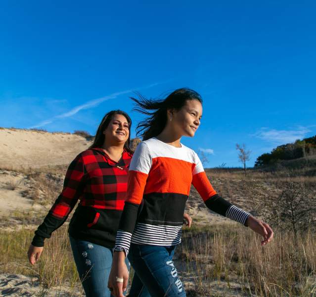 A woman and teen hike in the dunes. The bright blue sky is behind them. Vegetation on dunes covers the ground.