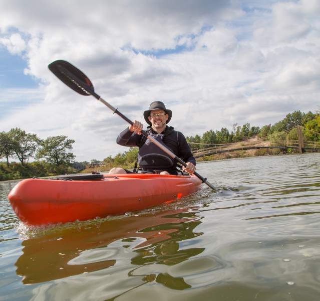 A man smiles as he sits in an orange kayak, paddling on the water. The sky is partly cloudy in the background.
