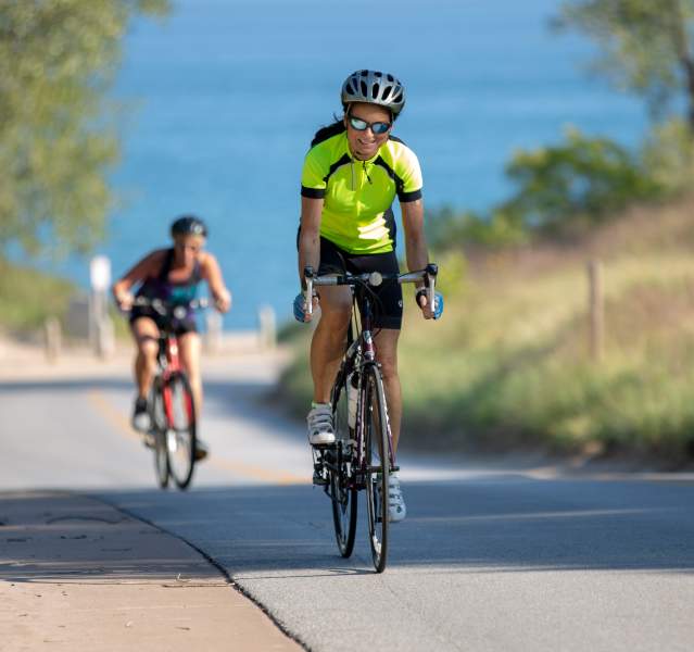 Bicyclists ride up hill on a paved road. A lake is in view in the background.