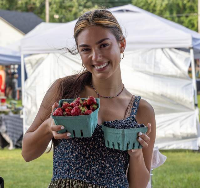 A woman holds two pints of berries. White tents are in the background.