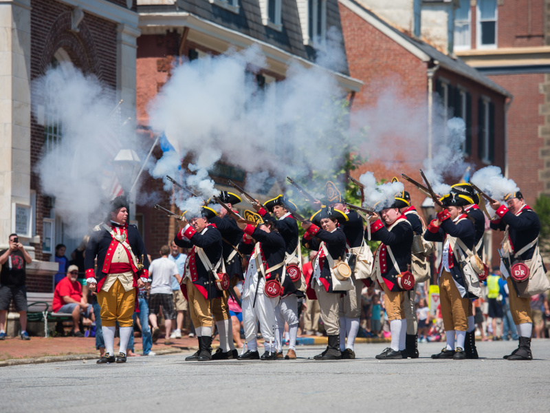 A group of historical reenactors in colonial military uniforms fires muskets, creating clouds of smoke during a lively outdoor event.