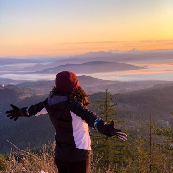 A person stands with their arms spread out on a snowy peak looking out over the foggy valley with sunset colors