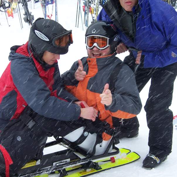 Two skiers help a skier strap onto an adaptive skiing board. They are on the snow and snow is falling.