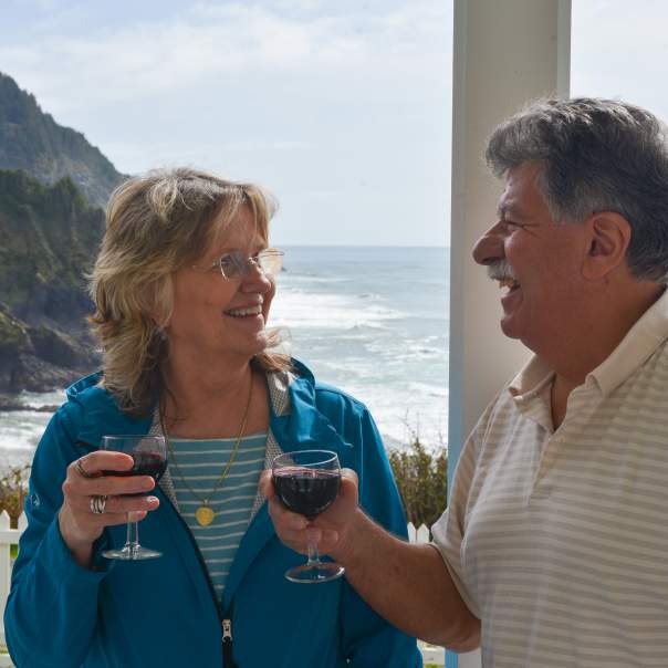 A couple drinking red wine is smiling with the ocean behind them.