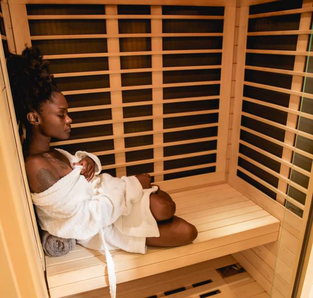 A black woman sits peacefully in a cedar sauna, wrapped in a white robe with eyes closed, enjoying a calming self-care moment in Fayetteville,