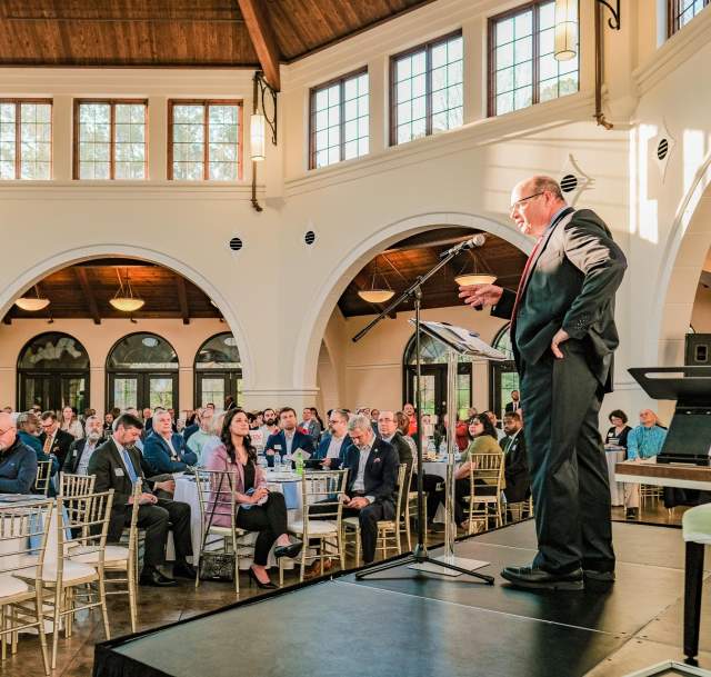 A professional speaker addresses a full room of attendees seated at banquet tables inside a bright, elegant event space at Cape Fear Botanical Garden in Fayetteville, North Carolina.